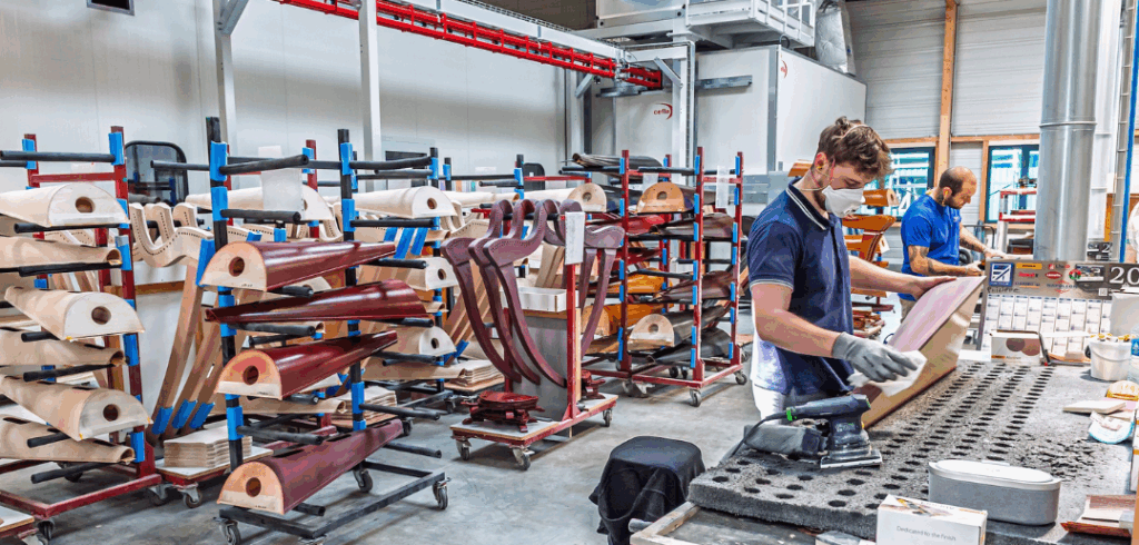Workers prepare wooden harp parts for assembly, with racks of more wooden harp parts in the background