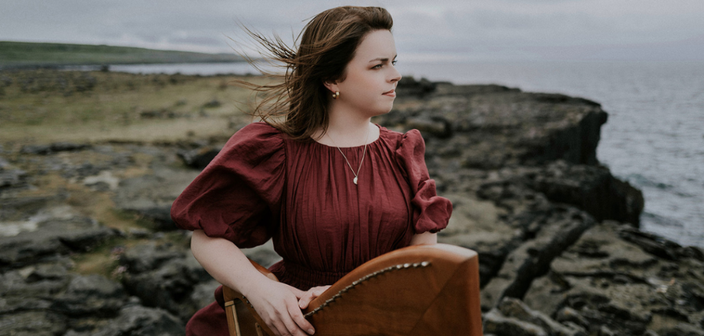 Aisling Lyons rests her hand on her Celtic harp with a rocky shoreline in the background