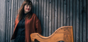 Rachel Hair stands against a weathered wooden wall beside her lever harp