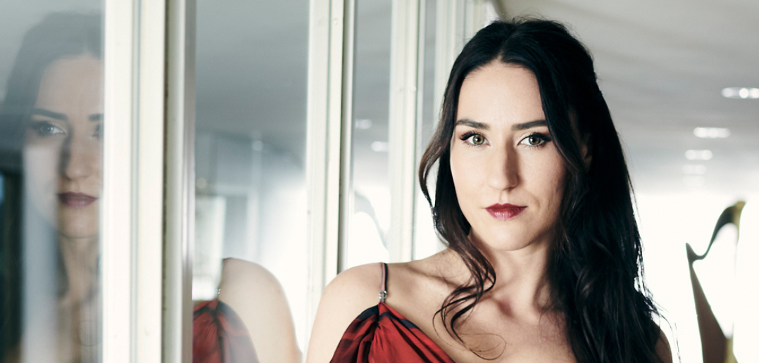 Clara_Bellegarde_pc_Matthias_Baus Clara Bellegarde stands in a hallway beside her reflection in glass windows, with a harp visible in the distance